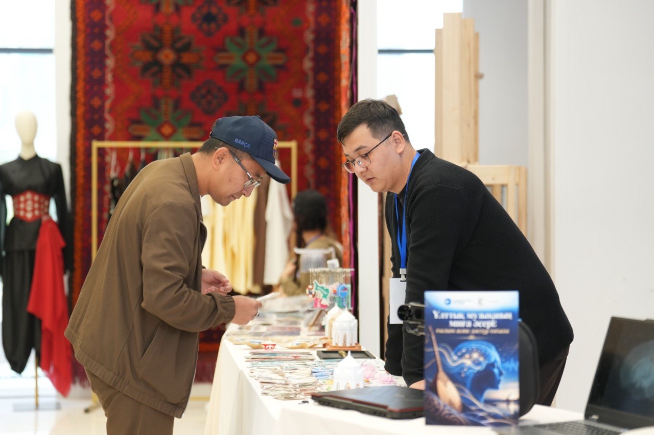 一名观众在阿特劳州集市的展台前观看展品，背景是带有纹样的地毯和服装。